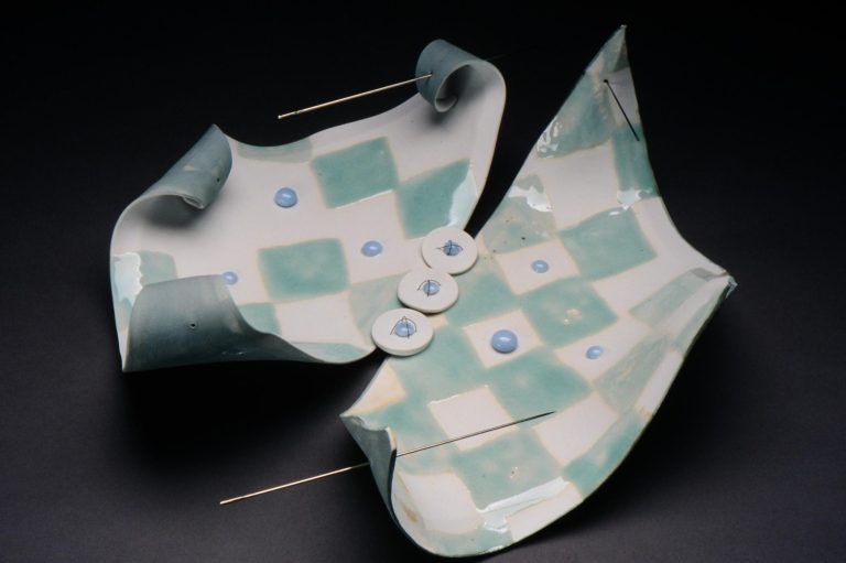 The image shows two wavy slabs of porcelain with a checkered celadon and white pattern. two long sewing needles puncture the slabs representing stitching.