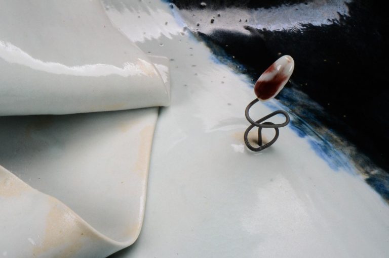 Close-up image of a wavy slab of porcelain in black and white with a red prescription pill on a wire that is stabbing the surface of bed sheets.