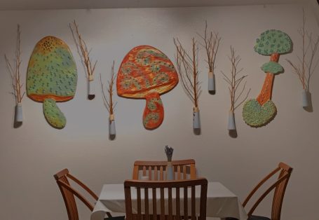 Wall installation, 9x3 feet long, of 3 mushrooms in bright green, orange, gold, and Kiln Jewels. Mixed media of plywood, and acrylic paint. 
