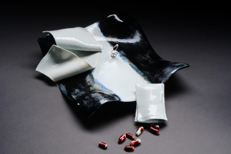 abletop sculpture of porcelain depicting disarrayed black and white sheets and pillows with red capsules spilling from a pillowcase.